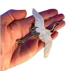 May include: White and black embroidered iron-on patch of a crane in flight. The crane has a long neck and long legs. The patch is perfect for adding a touch of nature to your clothing or accessories.