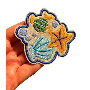 May include: Embroidered patch depicting a beach scene with a green and yellow striped fish, a yellow starfish with a purple centre, and a blue seashell. The patch has a blue border and is on a sandy background.