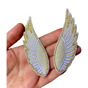 Può includere: Due toppe ricamate a forma di ali. Ogni ala presenta un centro grigio chiaro, bordato da cuciture azzurre e rifiniture dorate. Le toppe sono progettate per essere cucite sul tessuto.