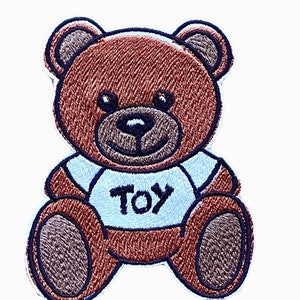 Könnte beinhalten: Brauner gestickter Teddybär, der ein weißes Hemd mit dem Wort "TOY" in schwarzen Buchstaben trägt.