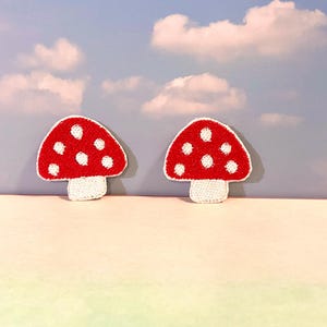 May include: Two red mushroom patches with white polka dots and white stems. The patches are embroidered and have a textured appearance. The background features a sky with clouds.