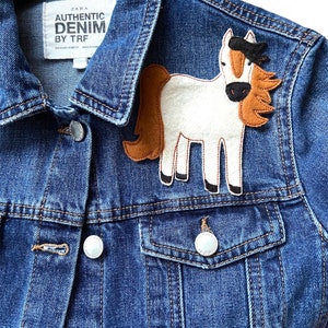 Può includere: Una giacca di jeans blu con un'applicazione di cavallo bianco sulla spalla sinistra. Il cavallo ha i capelli marroni e una criniera nera. La giacca ha un'etichetta che recita "ZARA AUTHENTIC DENIM BY TRF".
