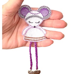Könnte beinhalten: Gesticktes Maus-Applikation in Weiß, Lila und Gold. Die Maus hat große, runde Ohren mit einem Kreuzschraffurmuster, geschlossene Augen und einen dekorativen Rock. Lila geflochtene Beine hängen von der Maus herab und enden in goldenen Herzanhängern.