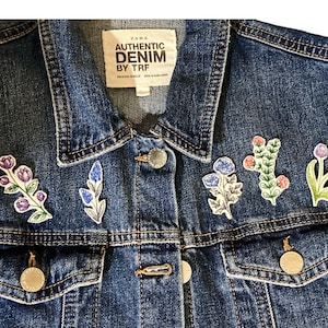 Puede incluir: Una chaqueta vaquera azul con cinco parches bordados de flores. Los parches son en tonos de azul, morado, rosa y verde. La chaqueta tiene una etiqueta que dice "ZARA AUTHENTIC DENIM BY TRF".