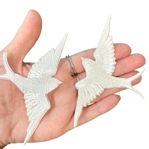 Puede incluir: Dos golondrinas blancas bordadas con las alas extendidas. Las aves están detalladas con costuras texturizadas y se sostienen en una mano. Las golondrinas están conectadas por una pequeña cadena.
