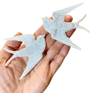 Puede incluir: Dos parches bordados de golondrina blanca. Los parches se muestran en una mano, con los pájaros mirando en direcciones opuestas.