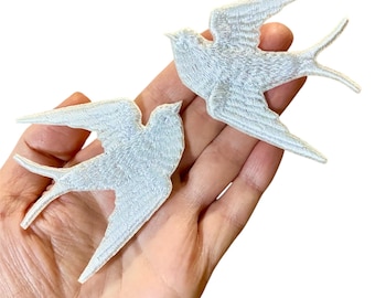 May include: Two white embroidered swallow bird patches. The patches are shown on a hand, with the birds facing opposite directions.