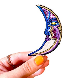 May include: A crescent moon shaped embroidered patch with a colorful face design. The face is outlined in gold thread and has a purple, blue, and yellow color scheme.