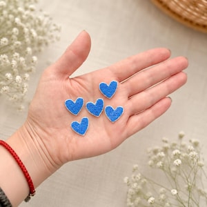 May include: Six small, blue heart-shaped felt appliques with white edges are displayed in an open hand. The hearts are a vibrant blue color and have a textured appearance. The background includes white flowers and a woven basket.