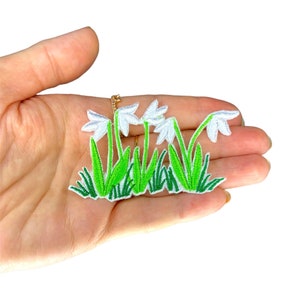 May include: White embroidered snowdrop flowers with green stems and leaves. The flowers are arranged in a row and are set against a white background.