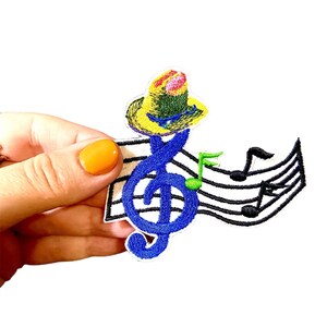 May include: An embroidered iron-on patch featuring a blue treble clef wearing a yellow and green hat, with a green musical note and black musical notes on a white background.