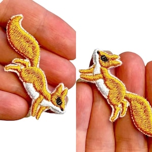 May include: Two embroidered iron-on patches of a yellow fox with white accents. The fox is in a jumping pose with its tail curled up.