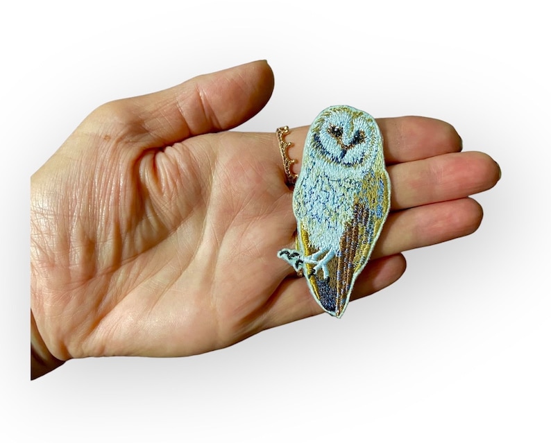 May include: A white and brown embroidered iron-on patch of a barn owl with a light blue background.