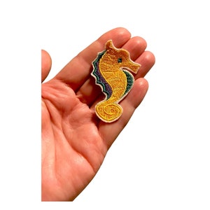 Könnte beinhalten: Gestickter Seepferdchen-Aufnäher in Gold-, Grün-, Lila- und Weißtönen. Das Seepferdchen wird in einer Hand gehalten, wodurch die detaillierte Gestaltung hervorgehoben wird. Der Aufnäher ist wahrscheinlich für Kleidung oder Accessoires.