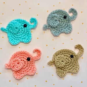 May include: Four crocheted elephant appliques in different colours: light blue, light pink, beige, and grey. Each elephant has a black button eye and a curled trunk.