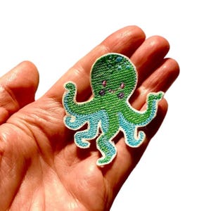Könnte beinhalten: Gestickter Oktopus-Aufnäher in Grün und Blau. Der Oktopus hat ein lächelndes Gesicht mit rosa Akzenten. Der Aufnäher befindet sich auf weißem Hintergrund. Ideal zum Aufnähen auf Kleidung, Taschen oder andere Stoffartikel.