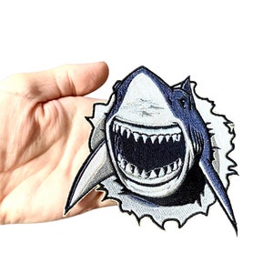 Peut inclure: Un patch brodé représentant un requin blanc et bleu avec la gueule ouverte, montrant des dents acérées. Le requin est représenté comme s'il jaillissait d'une surface, les bords du patch étant effilochés pour donner l'impression d'une déchirure.