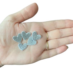May include: Five small, silver embroidered heart patches. The hearts are made of a textured, shimmering fabric. They are arranged in a loose cluster. These patches are suitable for adding a decorative touch to clothing or accessories.