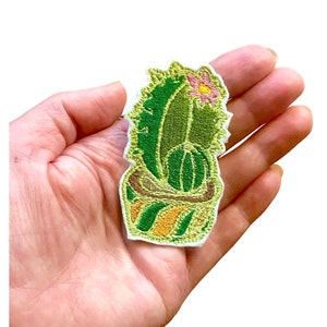 Puede incluir: Un parche bordado de cactus verde con una flor rosa en la parte superior, en una maceta a rayas amarilla y verde. El parche es termoadhesivo y se puede utilizar para decorar ropa, bolsos u otros artículos.