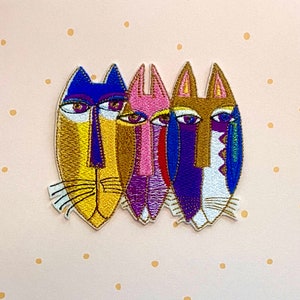 Könnte beinhalten: Vier bunte gestickte Aufnäher mit stilisierten Tiergesichtern. Die Aufnäher sind in Gold-, Rosa-, Lila-, Blau- und Weißtönen gehalten.