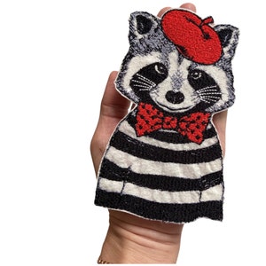 Könnte beinhalten: Gestickter Bügel-Patch mit einem Waschbären, der eine rote Baskenmütze und eine rote Schleife mit weißen Punkten trägt. Der Waschbär trägt ein schwarz-weiß gestreiftes Hemd.