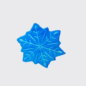 Puede incluir: Adorno azul en forma de copo de nieve con un diseño detallado y texturizado. El copo de nieve tiene seis puntas y un patrón simétrico de líneas que irradian desde el centro. El adorno es de color azul brillante y vibrante.
