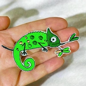 Puede incluir: Parche verde bordado de un camaleón de dibujos animados con una lengua larga y una hoja verde en la boca.