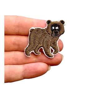 May include: Brown embroidered bear patch with white outline. The bear is standing on all fours and has a brown nose and black eyes.