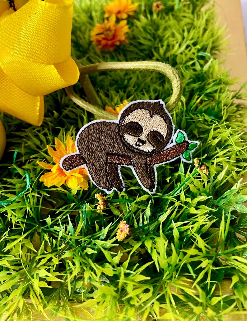 K&ouml;nnte beinhalten: Ein braunes Faultier mit geschlossenen Augen ist auf einem gr&uuml;nen Stoffhintergrund gestickt. Das Faultier h&auml;lt sich an einem Ast mit gr&uuml;nen Bl&auml;ttern fest.