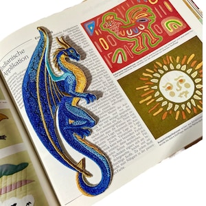 May include: A blue and gold embroidered dragon applique patch. The dragon is curled up with its tail in its mouth. The patch is on a book with text about Indian applique.