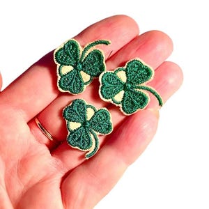 May include: Three green four-leaf clover patches with white centers and yellow stitching. The patches are embroidered and have a stem.