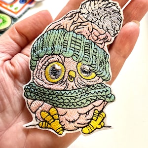 Op de afbeelding: Geborduurde patch van een uil met een groene gebreide muts en sjaal, met een bijpassende pompon. De uil heeft gele ogen en poten en een roze lichaam. Andere patches zijn zichtbaar op de achtergrond.