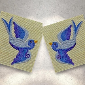 Puede incluir: Dos pájaros azules bordados con picos amarillos sobre un fondo crema. Los pájaros están mirando en direcciones opuestas y tienen un degradado de morado a azul.