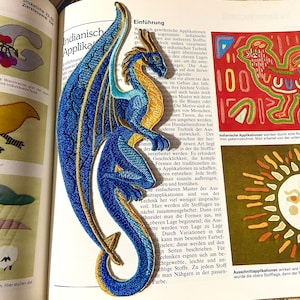 Könnte beinhalten: Ein gestickter Drachen-Applikations-Patch in Blau und Gold. Der Drache blickt nach rechts und hat seine Flügel ausgebreitet. Der Patch befindet sich auf einer Seite eines Buches über indische Applikationen.