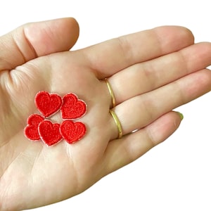 Può includere: Cinque toppe a forma di cuore in feltro rosso con cuciture bianche. Le toppe sono piccole e sono tenute in una mano.