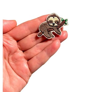 K&ouml;nnte beinhalten: Ein gestickter Faultier-Aufn&auml;her mit einem braunen Faultier, das einen schlafenden Ausdruck hat und einen gr&uuml;nen Zweig mit Bl&auml;ttern h&auml;lt. Der Aufn&auml;her ist auf wei&szlig;em Hintergrund.