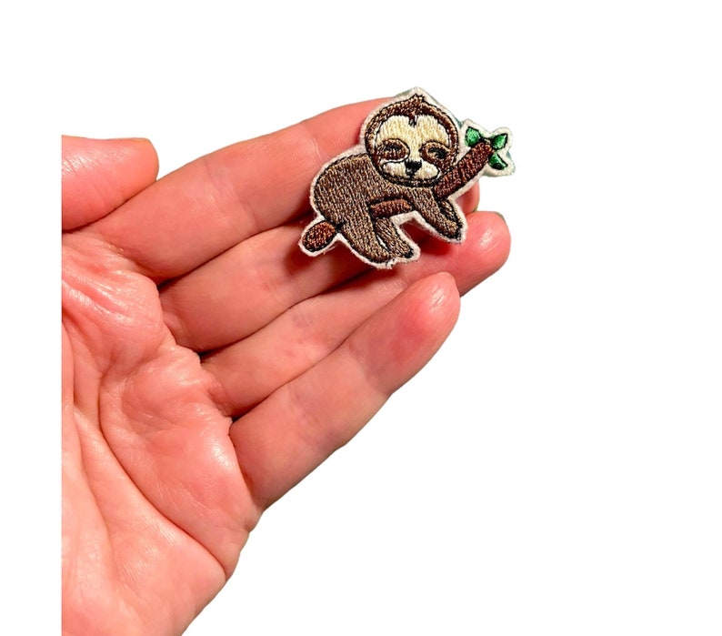 K&ouml;nnte beinhalten: Ein gestickter Faultier-Aufn&auml;her mit einem braunen Faultier, das einen schlafenden Ausdruck hat und einen gr&uuml;nen Zweig mit Bl&auml;ttern h&auml;lt. Der Aufn&auml;her ist auf wei&szlig;em Hintergrund.