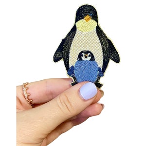 May include: Embroidered iron-on patch featuring a black and white penguin with a smaller blue penguin nestled inside. The larger penguin has a yellow beak and a yellow belly.
