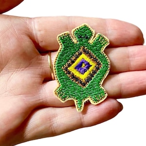 May include: Green embroidered turtle patch with a yellow, purple, and black diamond pattern in the center. The patch is on a beige background.