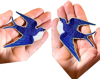 May include: Two blue and white embroidered swallow bird patches. The patches are on a hand, ready to be ironed on.