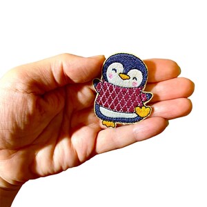 May include: A blue and white penguin wearing a red and white sweater with a diamond pattern. The penguin is smiling and has yellow feet.