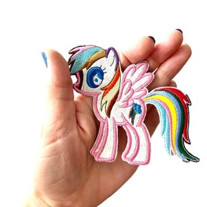 May include: Embroidered iron-on patch featuring a pink and white cartoon pony with a rainbow mane and tail. The pony has a blue eye and a pink flower on its flank.