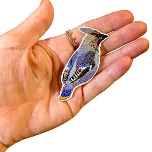 May include: A blue and brown embroidered iron-on patch of a bird with a white background.
