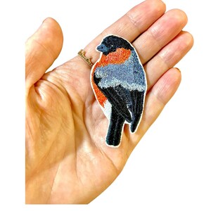 May include: Embroidered iron-on patch of a black, red, and blue bird. The bird is facing to the right and has a white belly.