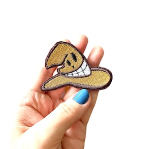 May include: Embroidered patch of a smiling cartoon character in a brown hat with a white brim and a brown border. The patch is held in a hand with blue nail polish. A fun, whimsical accessory.