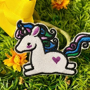 May include: Embroidered patch of a white unicorn with a black outline, colorful mane, and a purple heart. The patch is on a bed of green grass with yellow flowers and a yellow ribbon in the background.