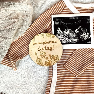 May include: A wooden round sign with the text "You are going to be a daddy!" in gold lettering. The sign is decorated with a floral design and is placed on a brown and white striped baby onesie. A black and white ultrasound image is visible in the background.