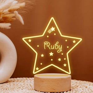 Könnte beinhalten: Ein hölzernes Nachtlicht in Form eines Sterns mit dem Namen "Ruby" im Inneren. Der Stern wird mit einem warmen gelben Licht beleuchtet und hat kleine Sterne, die um den Umfang verstreut sind.