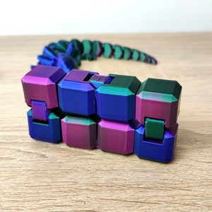 Può includere: Un giocattolo flessibile multicolore composto da cubi collegati. I cubi sono un gradiente di viola, blu e verde. Il giocattolo ha la forma di un serpente.