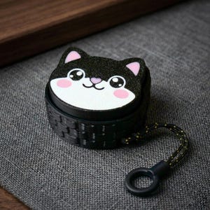 Könnte beinhalten: Schwarzes Katzen-Accessoire mit gewebter Textur. Das Katzengesicht hat rosa Ohren und Wangen sowie große Augen. Ein schwarzes und goldenes Schlüsselband mit Schlaufe ist befestigt.